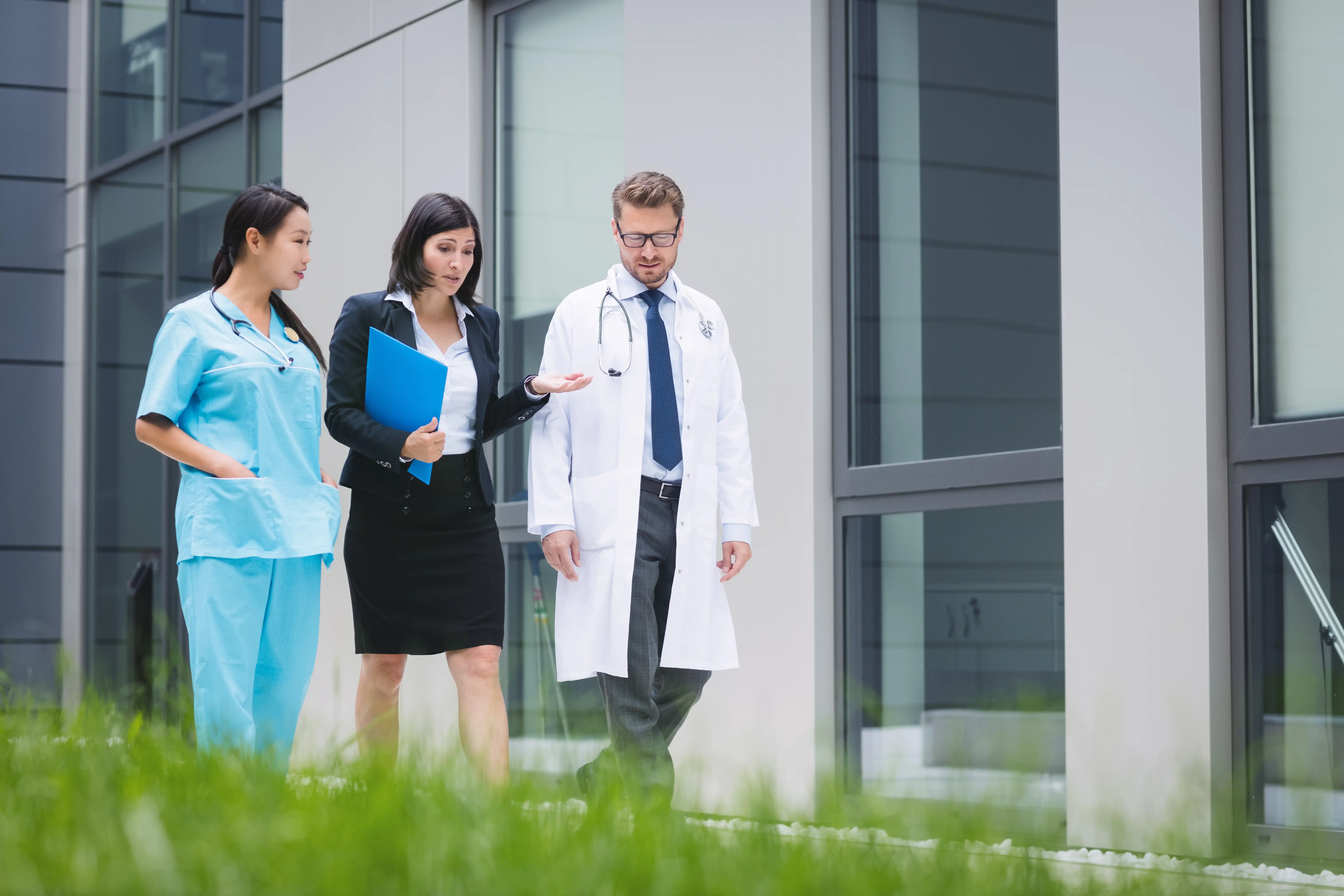 Three doctors walking together outside a building, engaged in conversation and wearing white coats.
