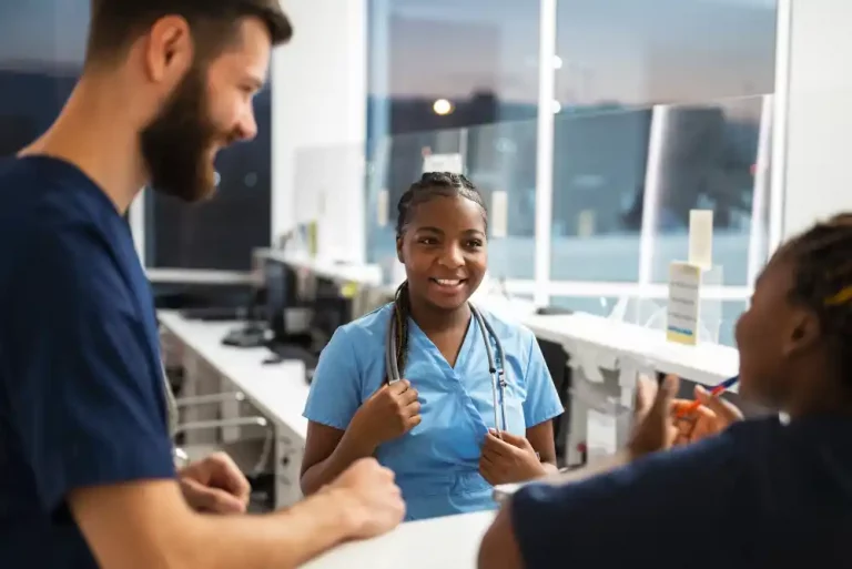 A group of healthcare professionals in scrubs converse and smile in a brightly lit hospital reception area, conveying a sense of teamwork and positivity.