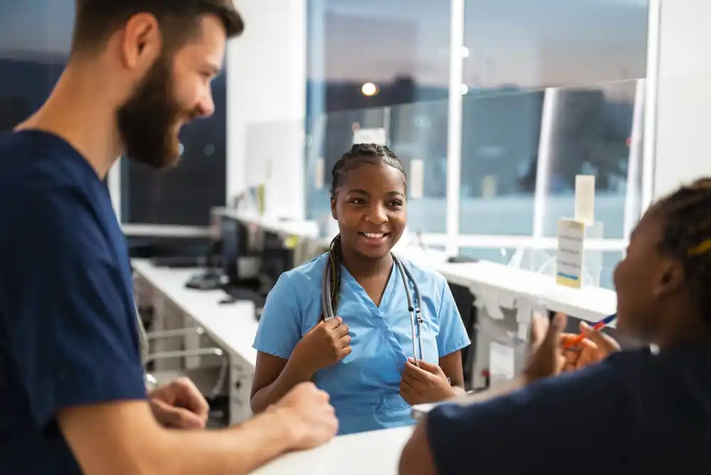 A group of healthcare professionals in scrubs converse and smile in a brightly lit hospital reception area, conveying a sense of teamwork and positivity.
