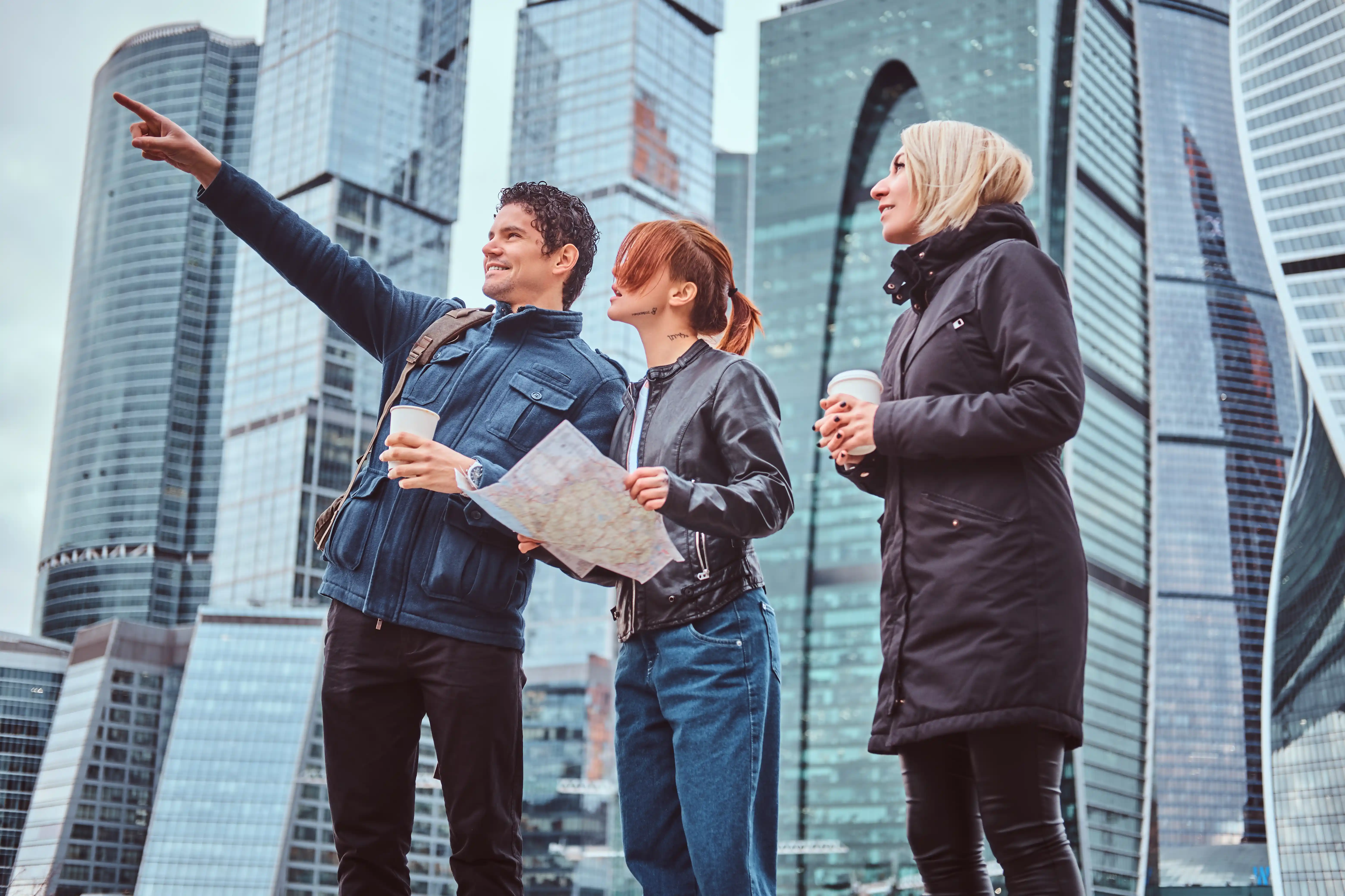 Three people stand outdoors in a cityscape, holding maps and coffee, with modern skyscrapers in the background. One person points forward, suggesting direction and exploration.