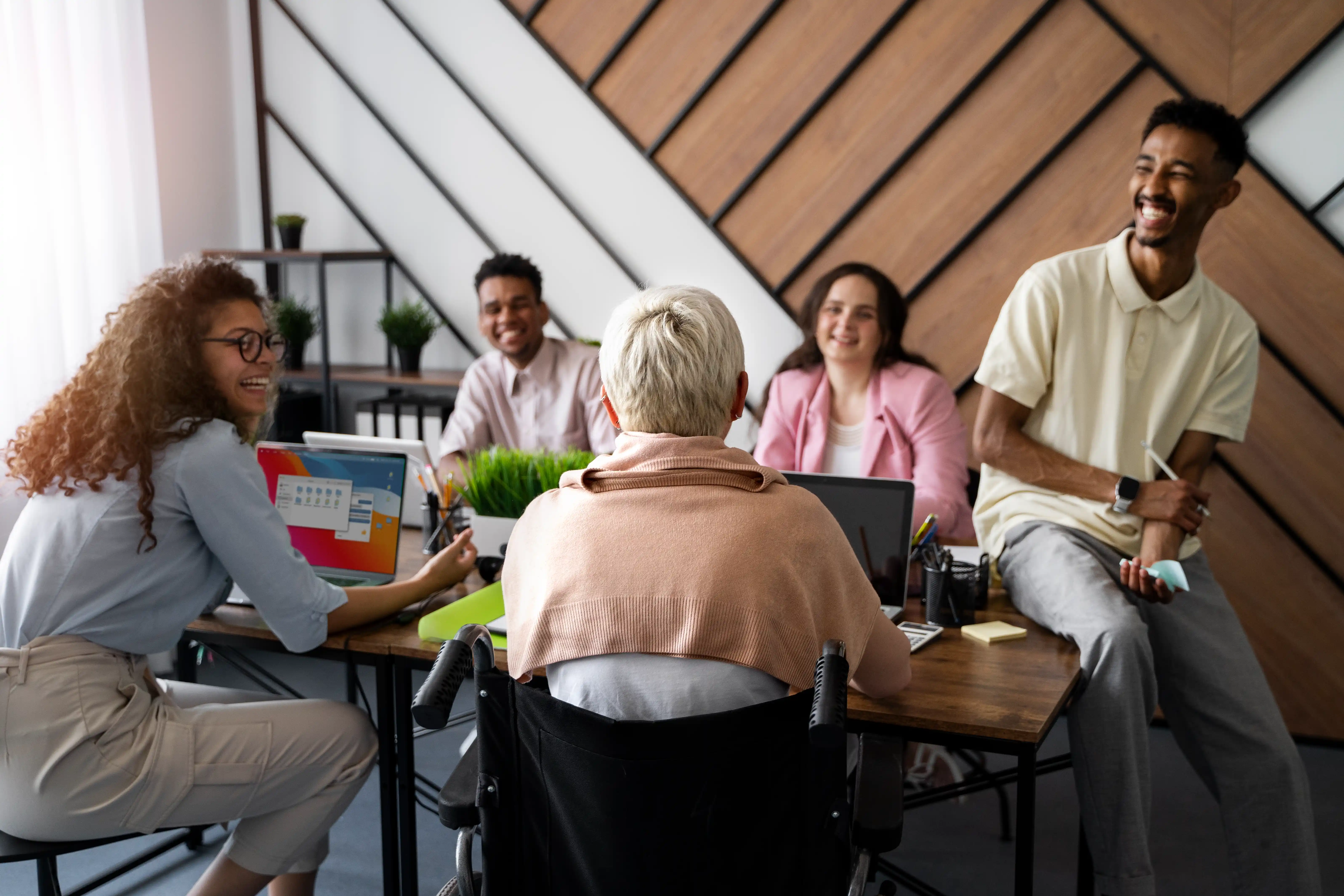 A diverse group of five people, including someone in a wheelchair, engage in a joyful team meeting around a table, with laptops and potted plants.