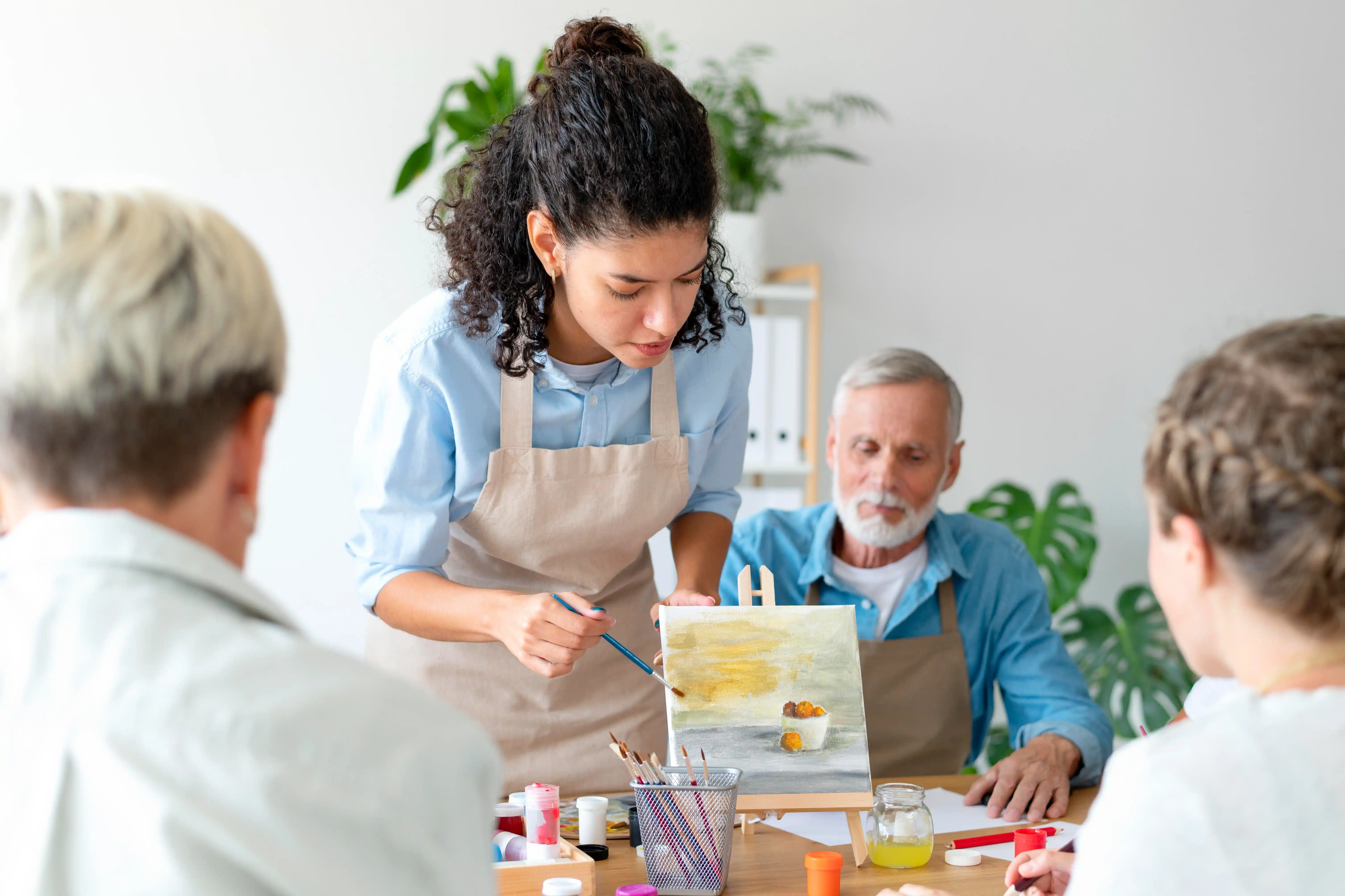 Artist teaches painting to a diverse group in a bright room. The scene feels calm and focused, as participants work on their canvases.