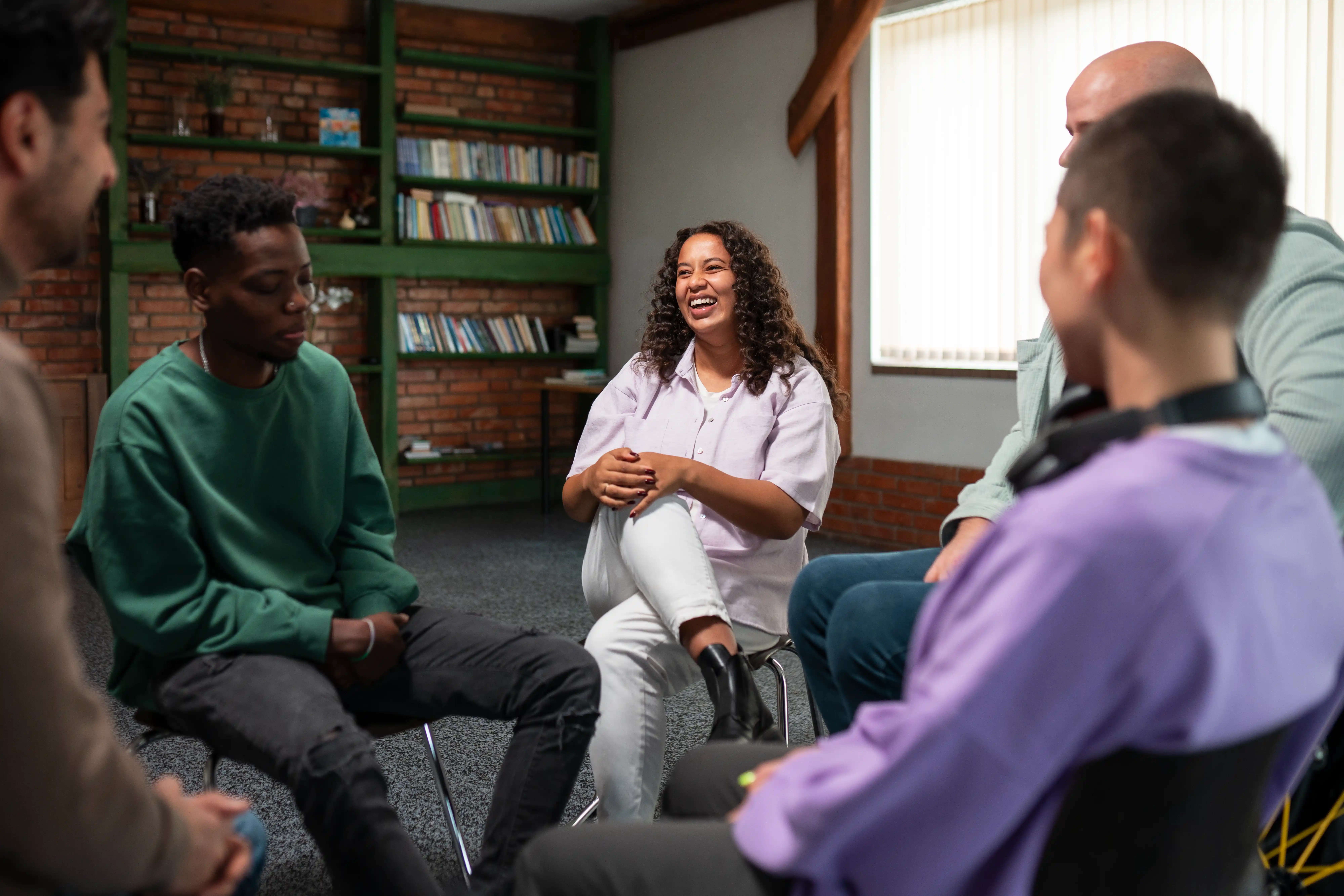 A group of five people sit in a circle, engaged in conversation, in a cozy room with bookshelves and large windows.